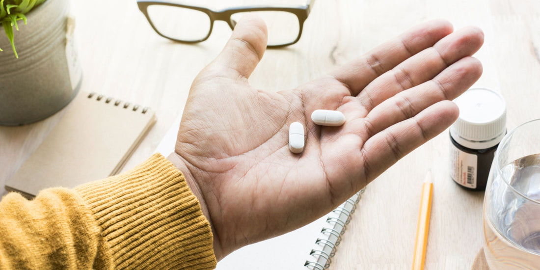 a person holding two white capsules, with a notepad, pencil, glasses, and a medication bottle nearby