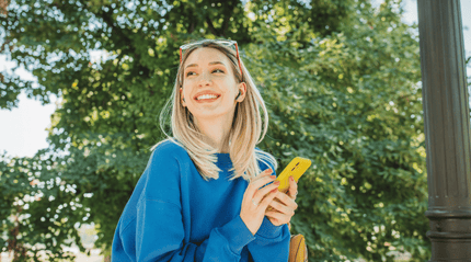 Person in a blue jumper holding a yellow phone outdoors in a leafy park setting. The bright, natural environment reflects everyday wellbeing and complements an article about supporting gut health with a simple daily probiotic routine.